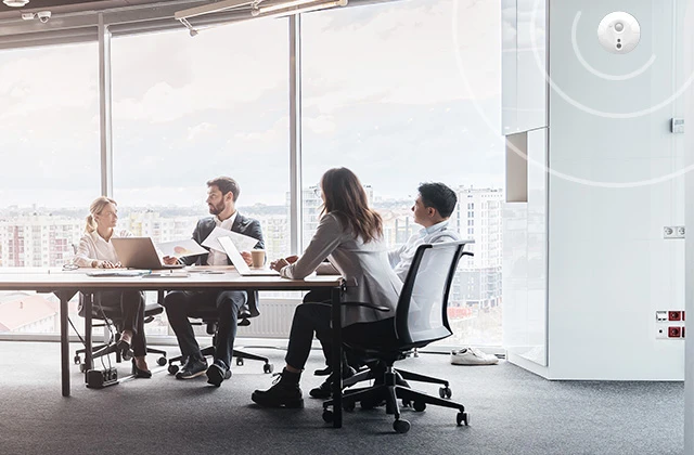 A wide shot of a contemporary office space with glass partitions, showing three people working at a table in the center. Sunlight streams in through large windows overlooking a cityscape.